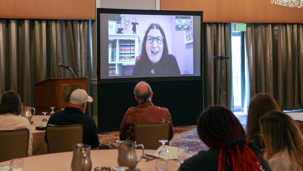 A screen at the front of a room shows Talia Swartz speaking with the backs of attendees in the foreground as they watch.