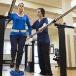 Woman being assisted by physical therapist, holding onto parallel bars and walking on a rubber balancing tool.