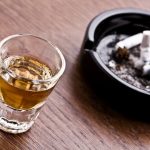 A shot glass of whiskey sits next to an ashtray with three discarded cigarettes.
