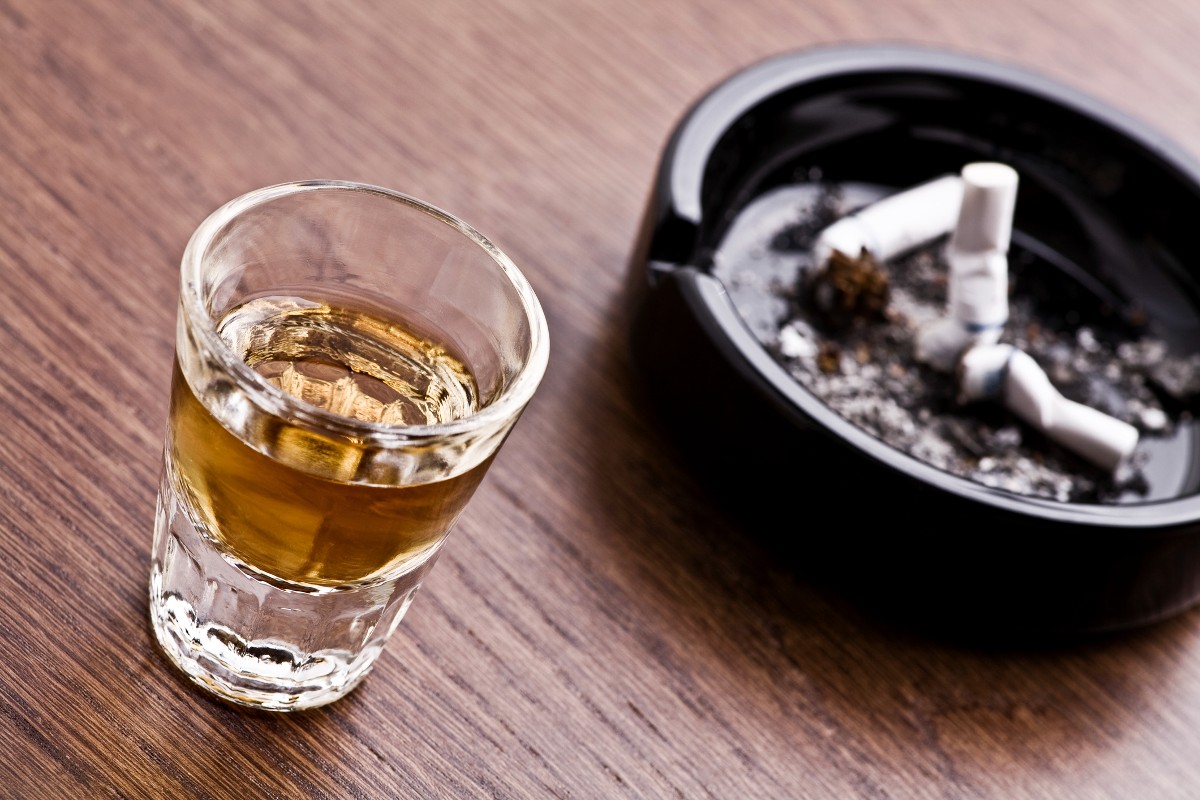 A shot glass of whiskey sits next to an ashtray with three discarded cigarettes.