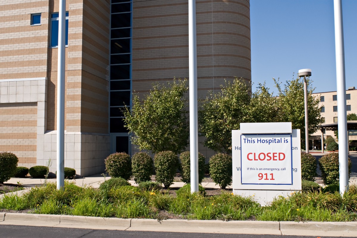 A hospital building sits vacant with a sign reading, “This Hospital is CLOSED. If this is an emergency, call 911.”