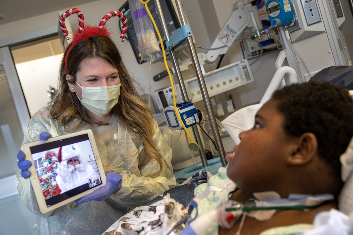 A woman in a candy cane headband, surgical mask and other personal protective equipment holds an iPad that bears a likeness of Santa Claus, while a young male patient looks on from his hospital bed.