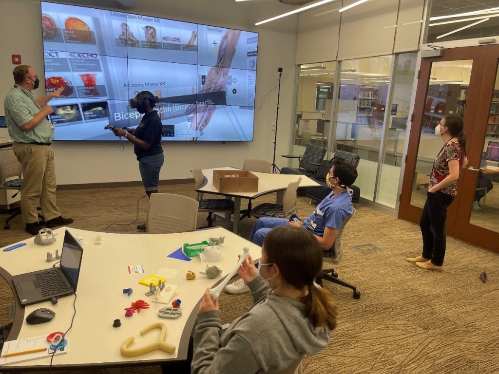 A man helps a student use virtual reality to study human anatomy while three other people watch. 