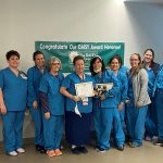 A group of women, most in hospital scrubs, pose for a photo. Two in the center hold plaques.