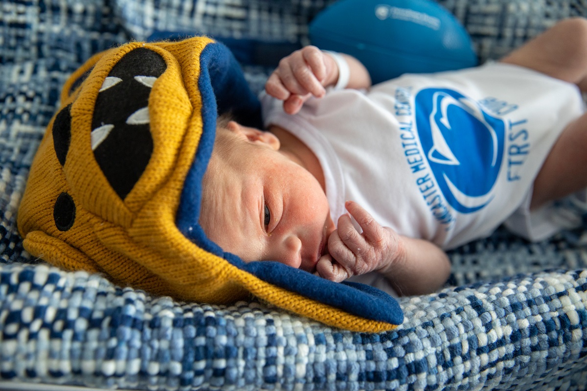 A baby lies on a cushion. He wears a Nittany Lion hat and a onesie with the Penn State logo.