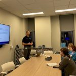 Four women sitting at a conference table looking at a man standing and talking in front of a TV screen showing a presentation slide.