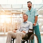 A young handsome doctor with his elderly disabled person in a wheelchair walk the corridors of a modern clinic.