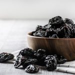 A bowl of prunes sits on a cloth on a countertop. Some prunes are also on the countertop.