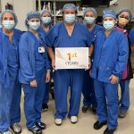 Seven people in surgical scrubs pose for a photo in a clinical space. The person in the middle holds a sign that reads “1st TCAR.”