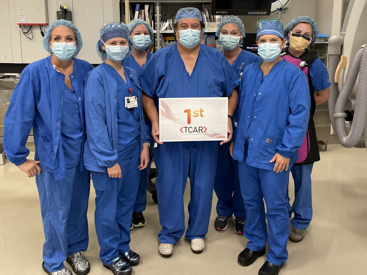 Seven people in surgical scrubs pose for a photo in a clinical space. The person in the middle holds a sign that reads “1st TCAR.”