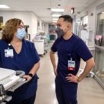 Arvin Ray Langurayan and Susan Tyson, both dressed in scrubs and wearing face masks, talk in the hallway outside a patient room at Holy Spirit Medical Center. Medical carts and equipment are in the background.