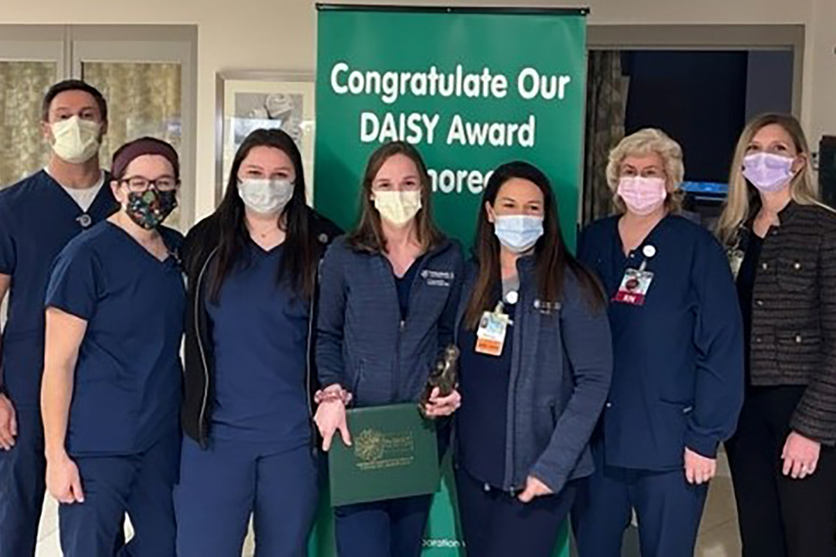 Seven people stand in a row. Six wear health care smocks. All wear surgical masks. The third woman from the left holds a statue and a plaque.