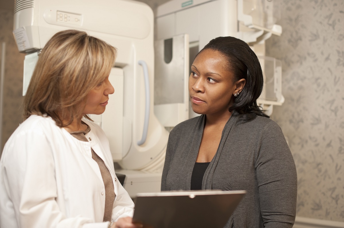 A technician explains a mammogram to an adult woman patient. A mammogram machine is in the background.