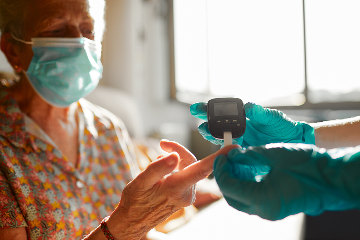 A masked woman holds her finger to a glucose meter that is held by another individual wearing gloves.
