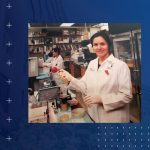 Joanna Floros stands at a bench in her lab while wearing a lab coat and holding a pipette.