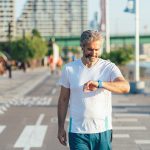 A middle-aged, bearded man wearing a short sleeve shirt walks along an urban waterfront while looking at his wearable device on his wrist.