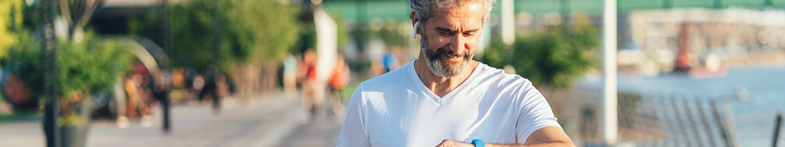A middle-aged, bearded man wearing a short sleeve shirt walks along an urban waterfront while looking at his wearable device on his wrist.