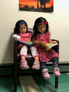 Two female children sharing an arm chair, each staring down at a book.