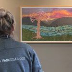 A woman facing away with a jacket that says, “Heart & Vascular CCU” on the back, views a mosaic artwork on a wall that depicts a flowering tree in front of a stream with hills and a sunrise in the background.