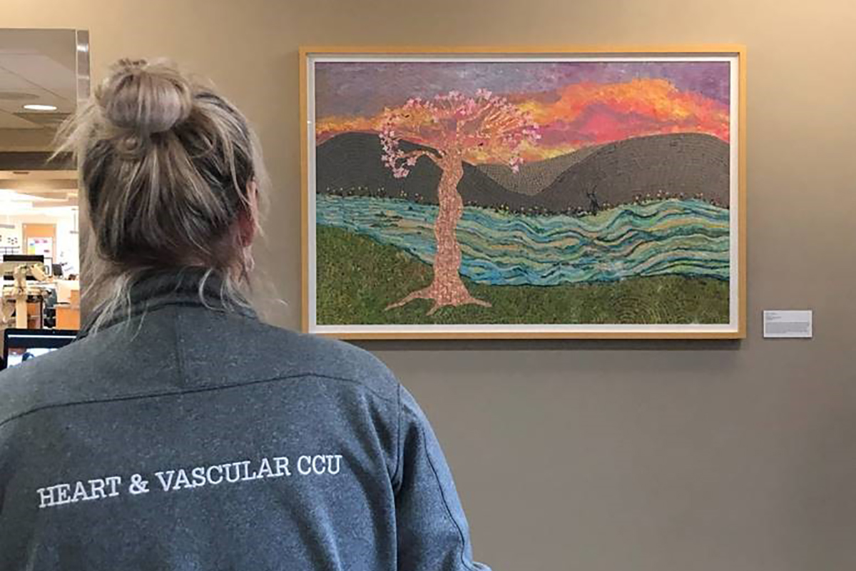 A woman facing away with a jacket that says, “Heart & Vascular CCU” on the back, views a mosaic artwork on a wall that depicts a flowering tree in front of a stream with hills and a sunrise in the background.
