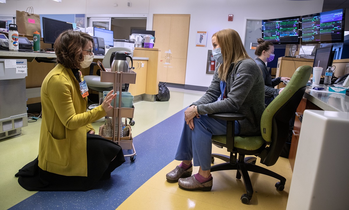 A woman in a cardigan sweater kneels in a hospital common room. She speaks with a woman in scrubs and a sweater sitting in a chair in front of her. Next to the kneeling woman is a rack of coffee pots and other implements. Behind the seated woman in a bank of computer screens, and another woman sits at a desk. Everyone is masked.