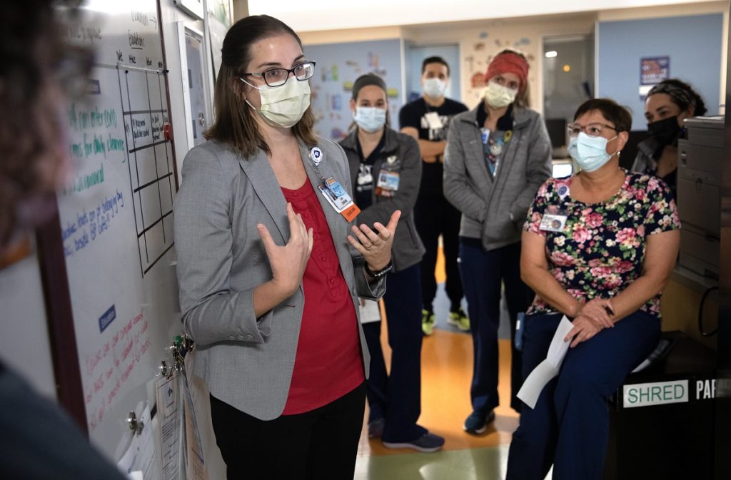 A woman in a mask stands against a wall in a hospital. She is surrounded by others in masks who listen to her as she speaks.