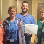 Five people stand in a row in a hospital room. The man in the center holds a certificate.