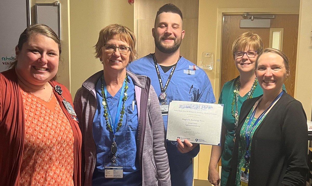 Five people stand in a row in a hospital room. The man in the center holds a certificate.