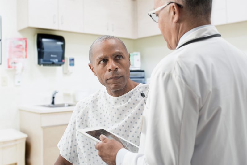 A doctor looks down at a tablet computer as he speaks with a male patient, who is seated in front of him.