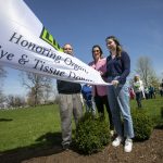 A man, a woman and a teenage girl hold a “Donate Life” flag while bystanders look on. Bushes and grass are in the foreground.