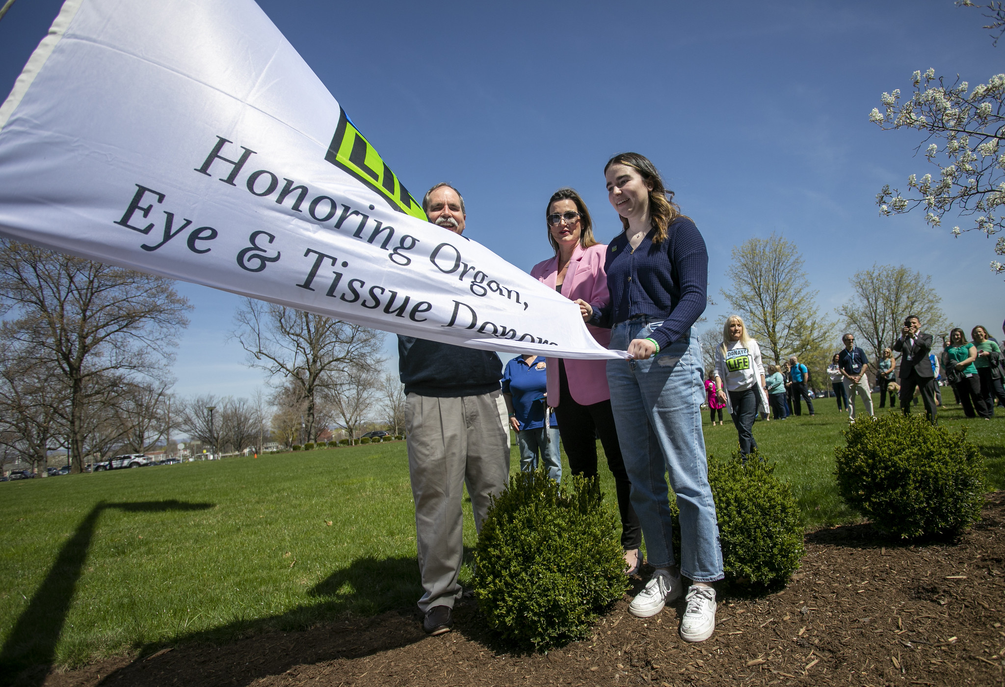 A man, a woman and a teenage girl hold a “Donate Life” flag while bystanders look on. Bushes and grass are in the foreground.