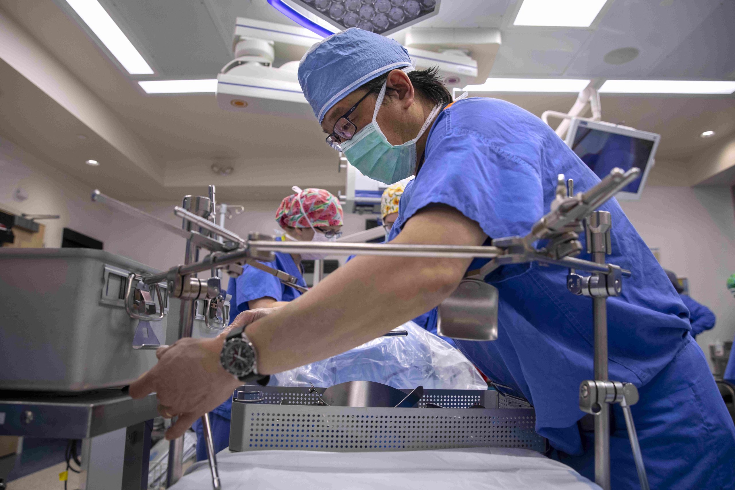 Dr. Johnny Hong, wearing surgical scrubs, works with a piece of equipment in the foreground of an operating room. Other staff members are working in the background.