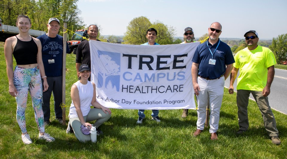 A group of volunteers gathers outdoors in the reforestation area of the Penn State Health Milton S. Hershey Medical Center campus around a sign that says “Tree Campus Healthcare: An Arbor Day Foundation Program.”