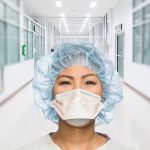 Head shot of female clinician wearing a surgical head cap and N95 mask, hospital doorway and hall in background.