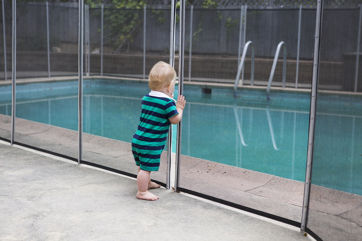 A toddler looks through a crack in a fence around a swimming pool.