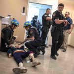 Two first responders attend to a mannequin – a simulated victim – while police officers and other drill participants look on.