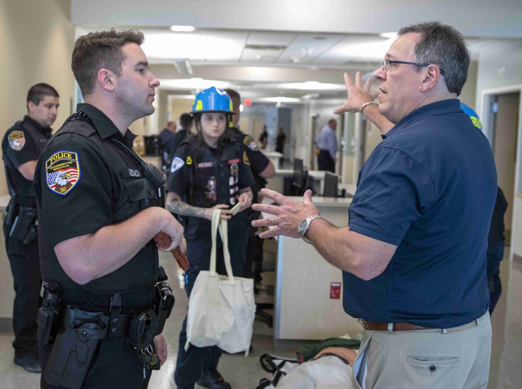 A man wearing a short-sleeved dress shirt talks with a police officer holding a fake handgun in a hospital hallway. Other officers are in the background.