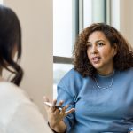Two women sitting and talking to each other. One faces the camera and gestures with her hand. The other faces away.