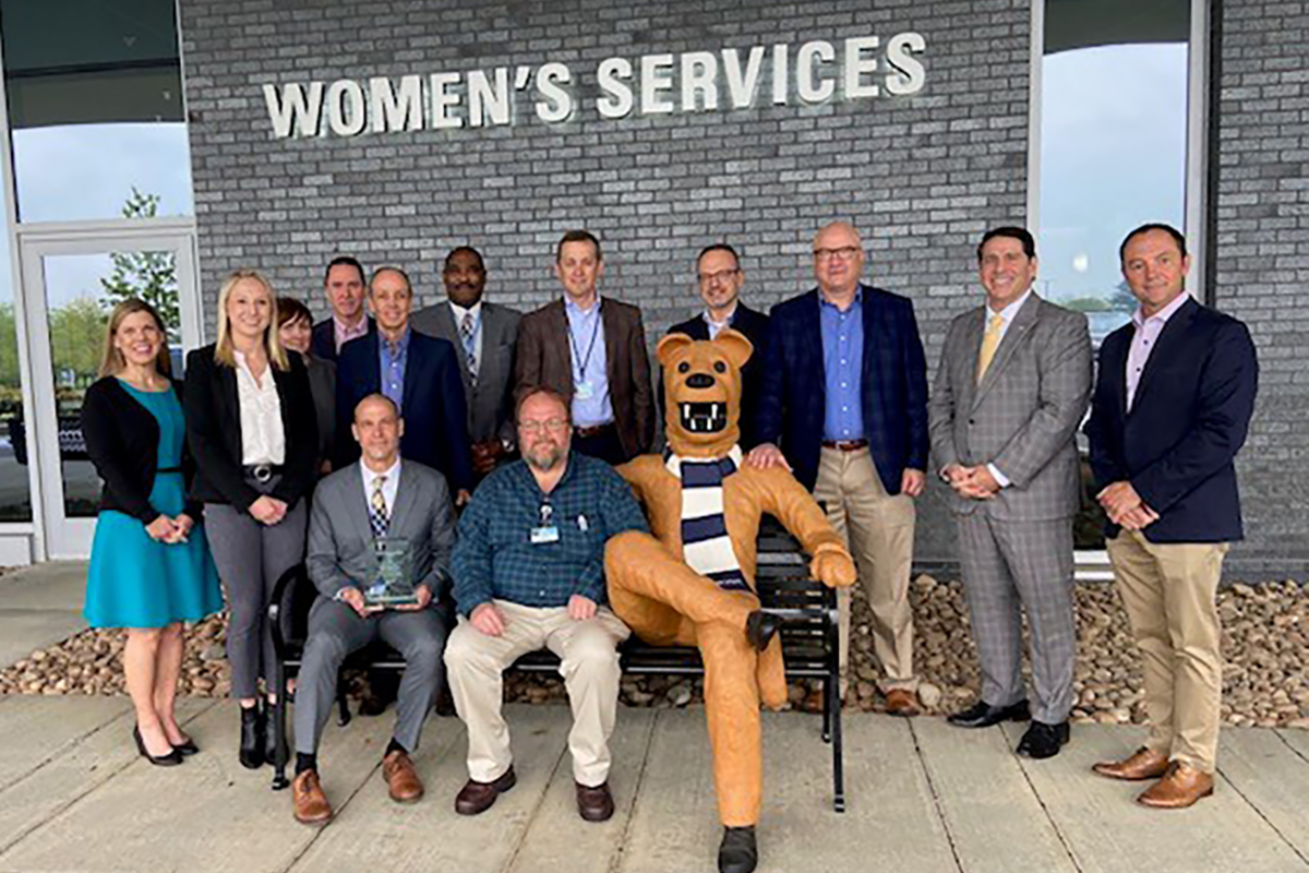 A man in a suit sits on a bench outside Penn State Health Hampden Medical Center holding a glass award. Another man in street clothes sits beside him. A permanently affixed life-size statue of the Penn State Nittany Lion is also seated on the bench. A large group of men and women including Penn State Health and FM Global leaders stand behind the bench.