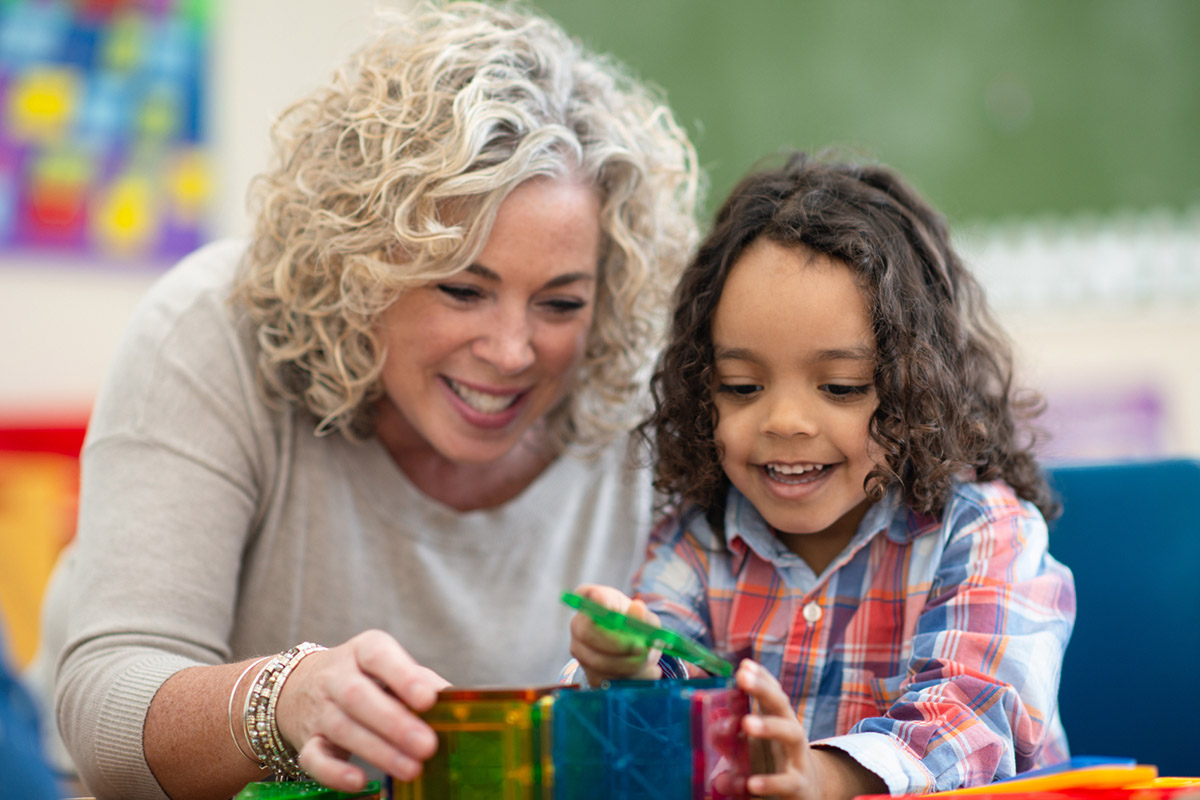 A teacher and child play with blocks in a classroom.