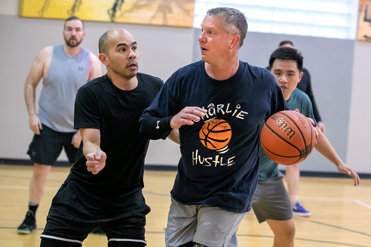 Man wearing athletic clothing dribbles basketball up a basketball court as other men chase him and look on.