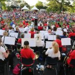 View from the back of an orchestra full of musicians performing before a crowd of a couple thousand people in an outdoor setting.