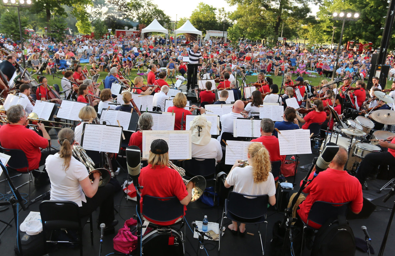 View from the back of an orchestra full of musicians performing before a crowd of a couple thousand people in an outdoor setting.