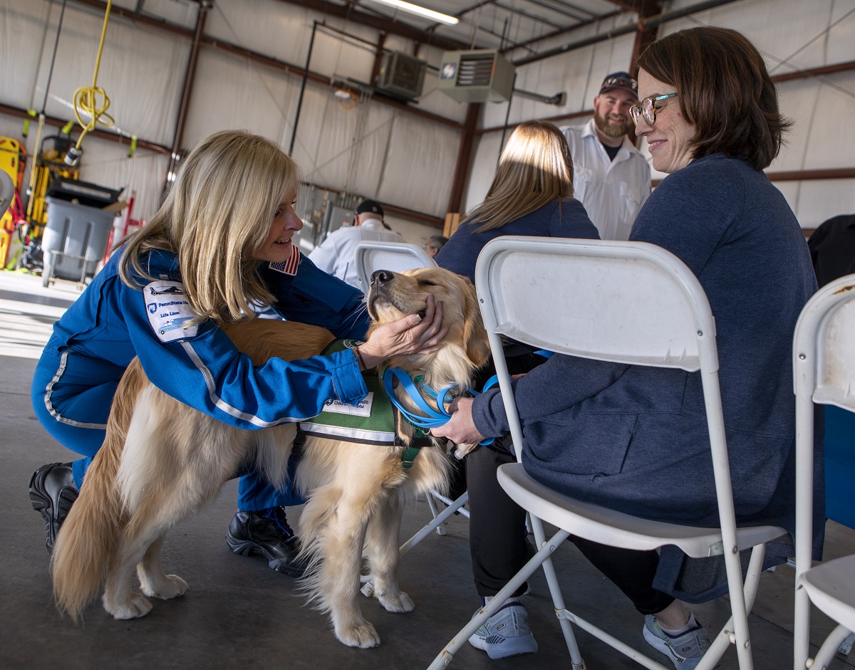 A woman pets a dog next to a woman seated in a chair.
