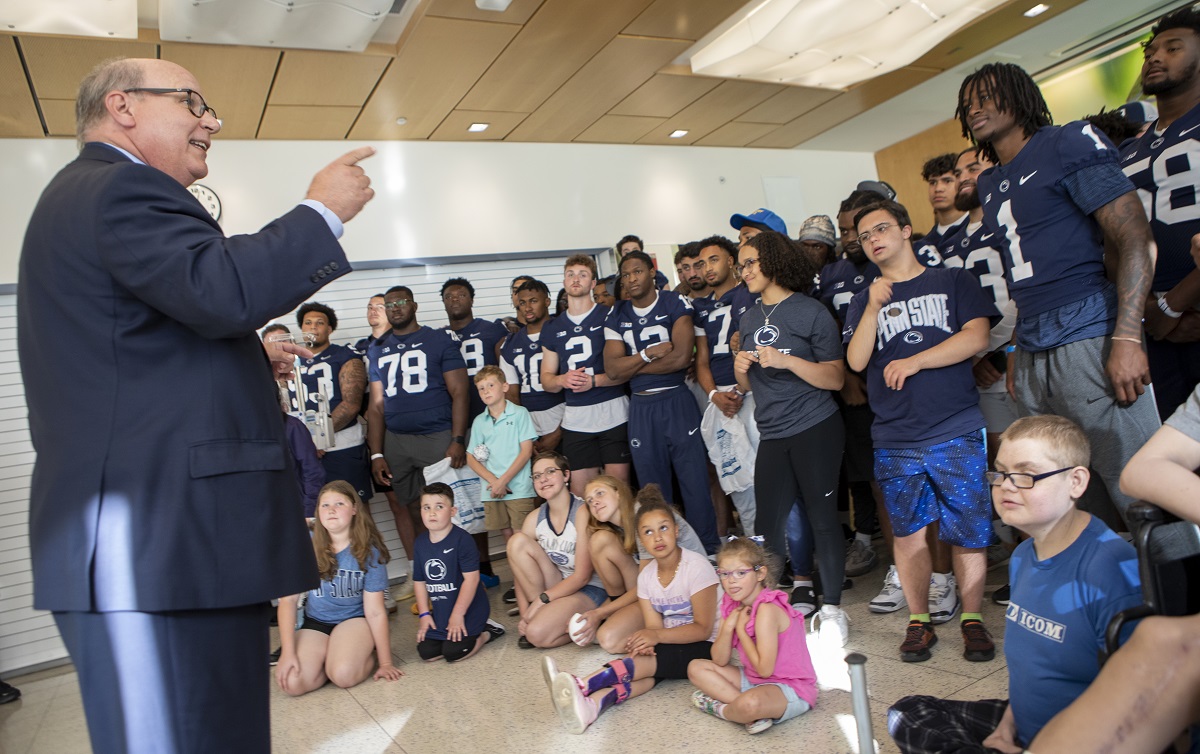 A man in a suit smiles and gestures at football players in their jerseys and a group of children.