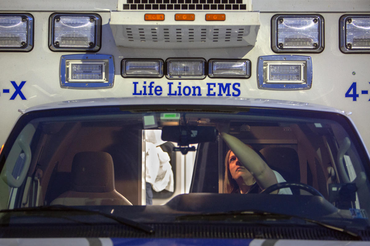A view through the windshield of an ambulance as a worker in the driver's seat reaches up to press buttons on the roof.