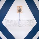 Photo shows a bell that pediatric cancer patients ring during their final cancer treatments at Penn State Health Children’s Hospital. The bell is places above a sign that says, “Conquer.”