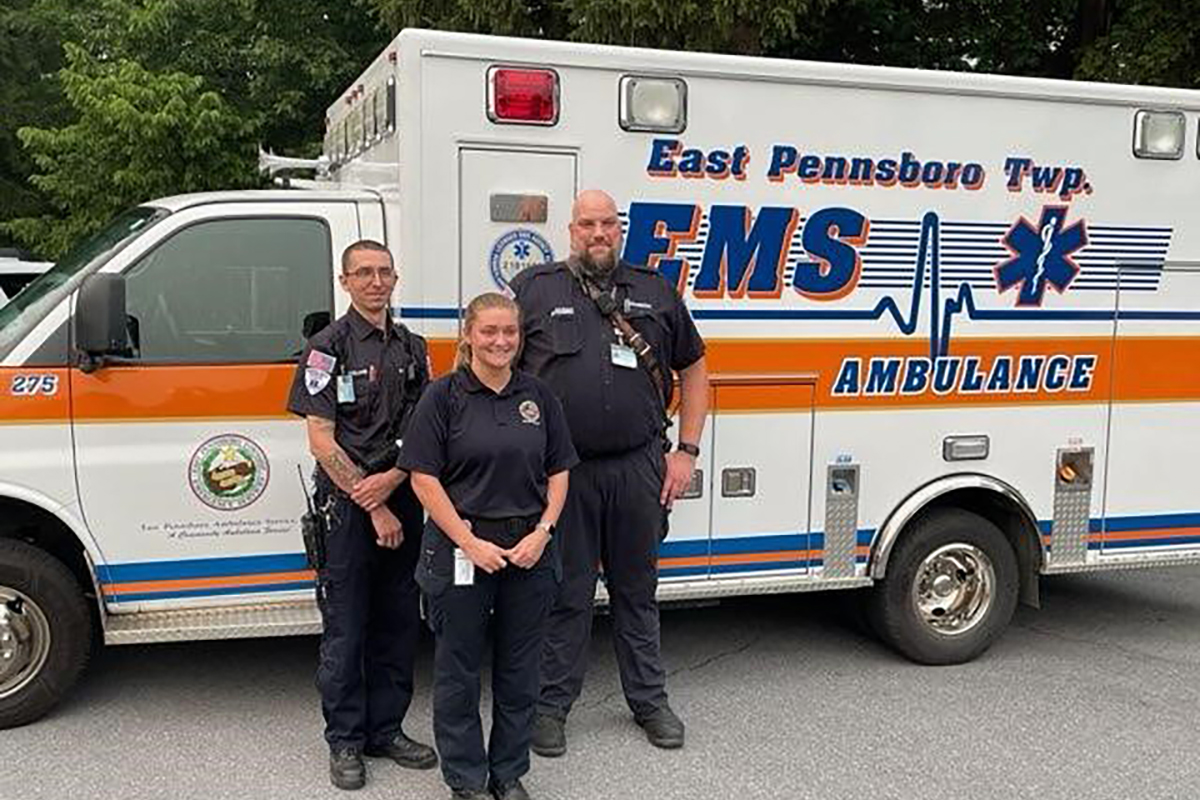 Three individuals in emergency medical personnel uniforms standing in front of an East Pennsboro Township ambulance.