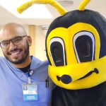 Gentleman wearing scrubs and smiling with his arm around a person in a bee costume.