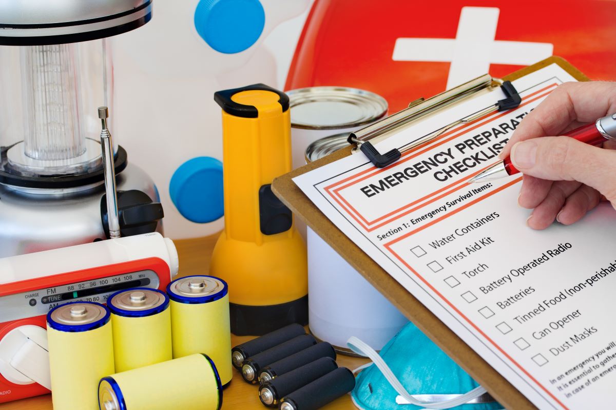 Clipboard with checklist titled “Emergency Preparation Checklist” – person’s hand hold pen over checklist. In the background: lantern, flashlight, batteries, first aid kit, mask, canned food, water jug, radio.
